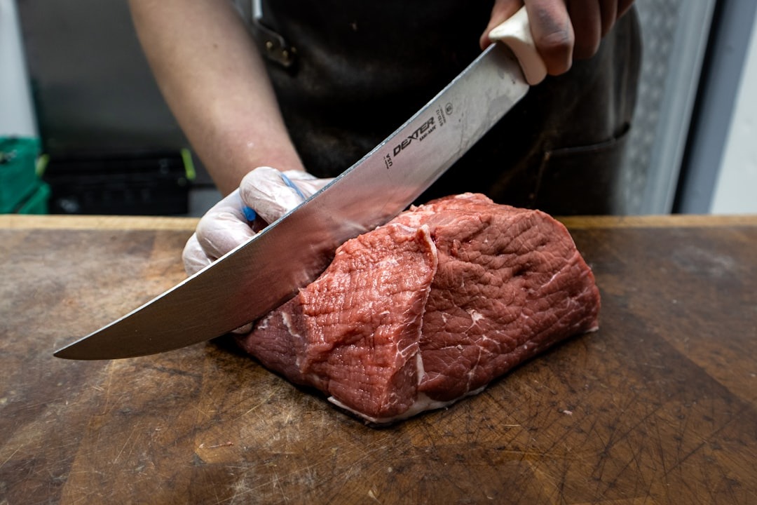 Gloved hands slice raw meat on a wooden cutting board.